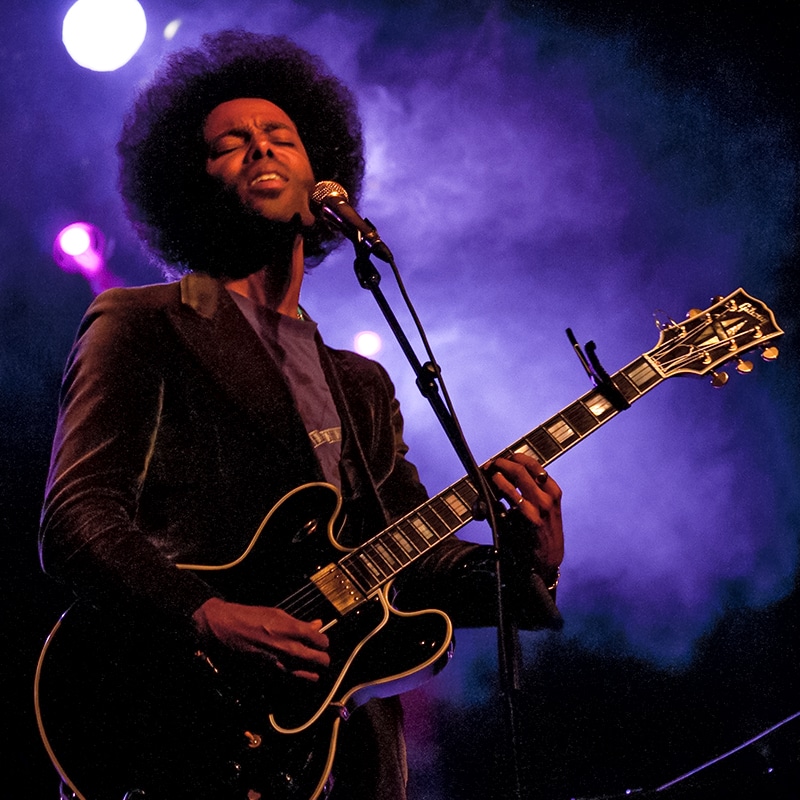 Concert photography, man playing guitar with a purple haze behind him.